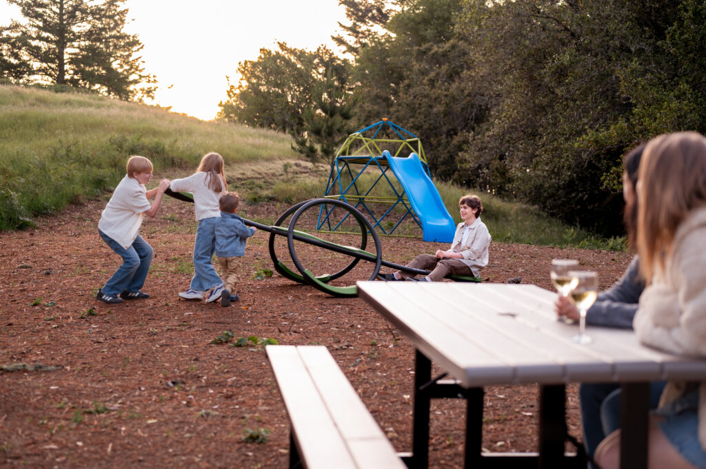 Four kids playing on teeter totter with slide in background, while two moms sit sipping wine on a picnic table.